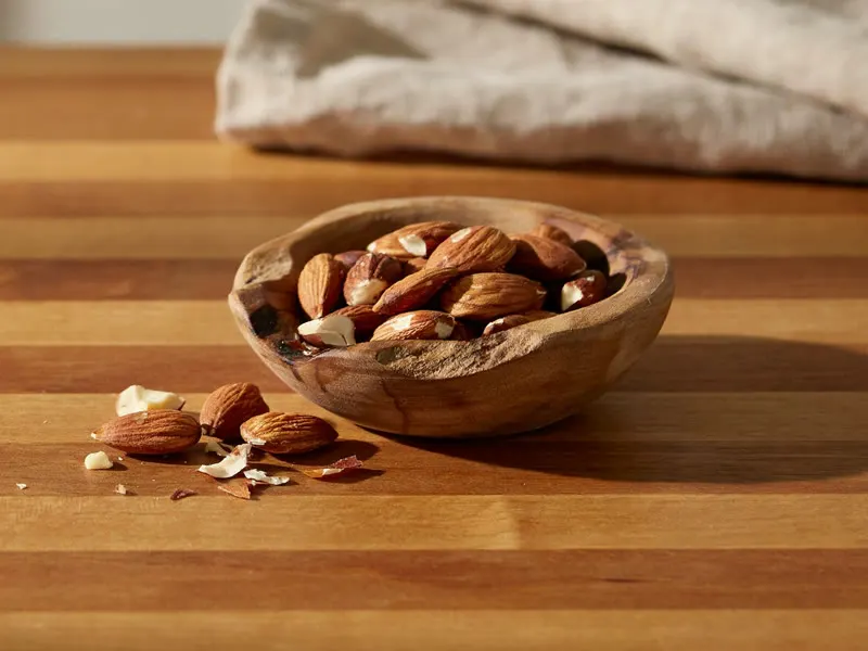 Raw almonds, some still in their shell, scattered on a wooden counter beside a small bowl of shelled almonds.