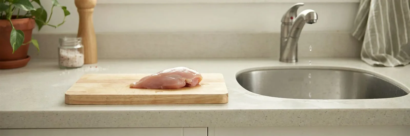 A raw chicken breast on a wooden cutting board on a kitchen counter, positioned close to a sink with the tap running.