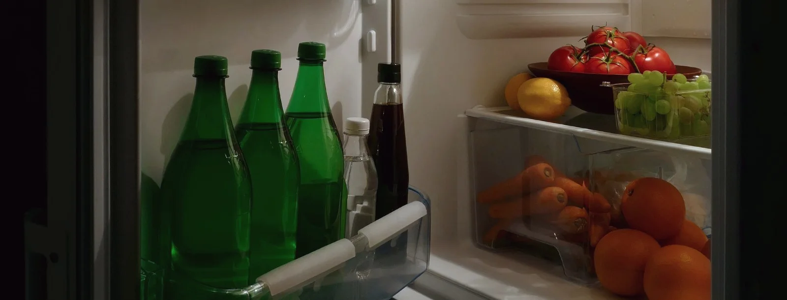 An open fridge at night, shelves holding tomatoes, grapes, carrots and oranges.
