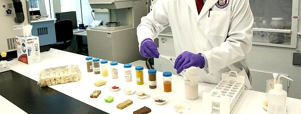 A lab technician in a white coat and purple gloves preparing food samples for analysis, sample vials lined up on the counter.